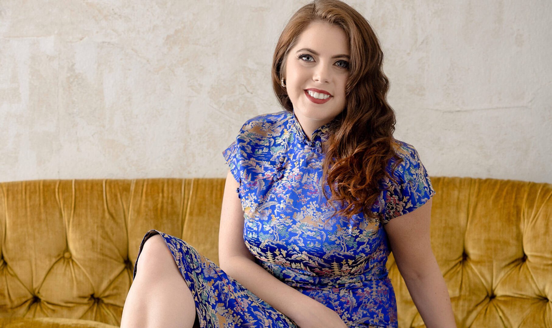 A woman in a blue floral dress sitting on a mustard couch, smiling.
